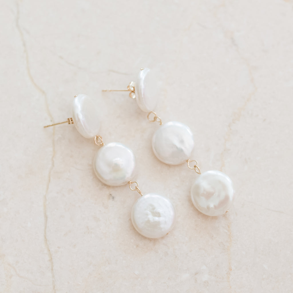 Triple Coin Pearl Earrings by Pearly Girls, a set of freshwater pearl earrings. These earrings feature three layers of coin pearls, known for their unique, flat shape, offering a modern twist on classic pearl earrings with a stacked, dimensional design that adds elegance and interest.