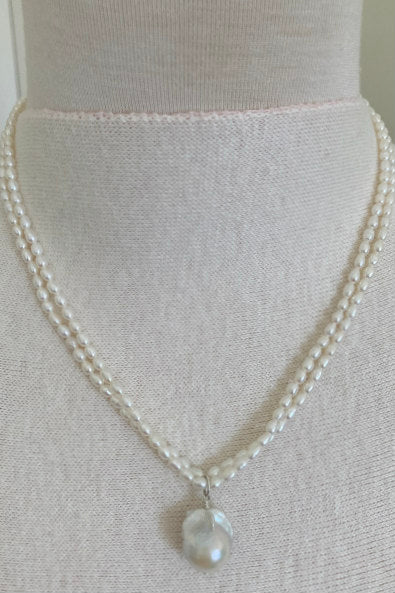 Claire Pearl Necklace by Pearly Girls features a fireball pearl pendant and seed pearls. This necklace elegantly showcases a striking fireball pearl pendant, complemented by a string of delicate seed pearls, creating a balance of boldness and subtlety in its design.